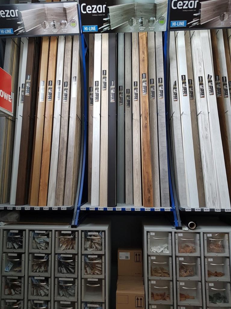 Ekspozycja różnorodnych paneli podłogowych marki Cezar Hi-Line w sklepie, ułożonych pionowo na stojakach, z widocznymi wzorami i kolorami, oraz szafkami z próbkami poniżej.