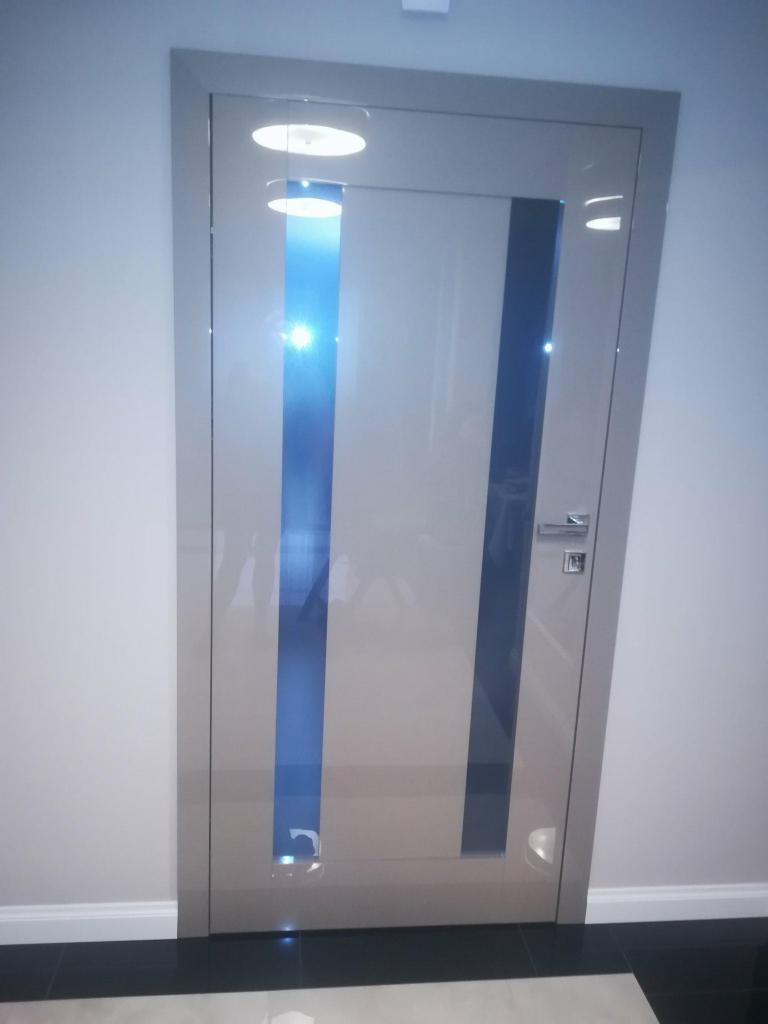 Szklane drzwi wewnętrzne z dwoma pionowymi, niebieskimi pasami, zamontowane w jasnym pomieszczeniu z czarno-białą podłogą.