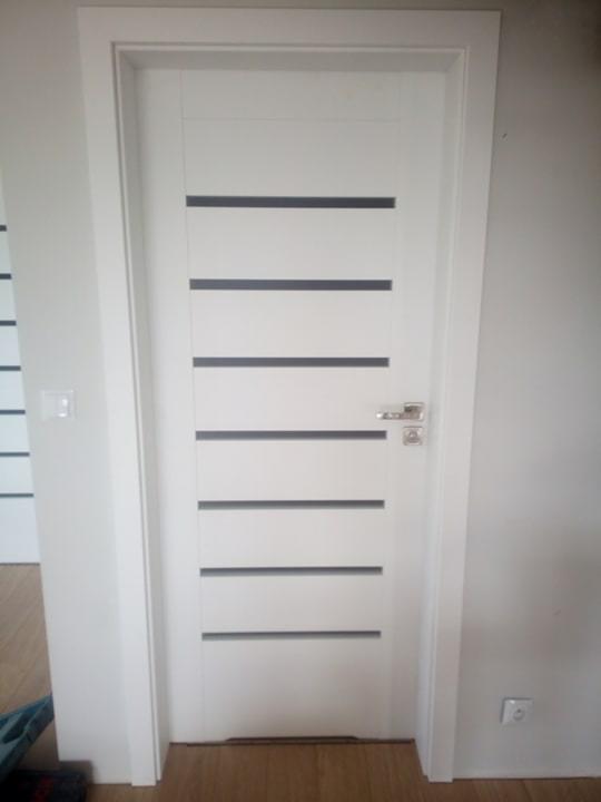 Białe drzwi wewnętrzne z poziomymi, szarymi wstawkami, zamontowane w ościeżnicy, widok na fragment podłogi i ściany z gniazdkiem.