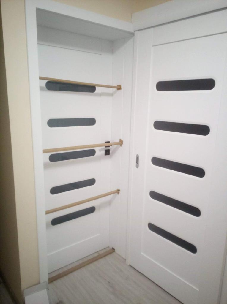 Białe drzwi wewnętrzne z poziomymi, owalnymi przeszkleniami, zamontowane w ościeżnicy; widoczne drewniane kołki w otworach po lewej stronie.