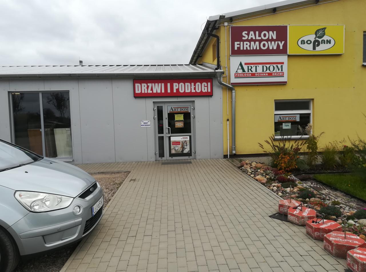 Fasada budynku firmy Art Dom z szyldem 'Drzwi i Podłogi', widoczny salon firmowy z żółtą elewacją, obok zaparkowany srebrny samochód osobowy.