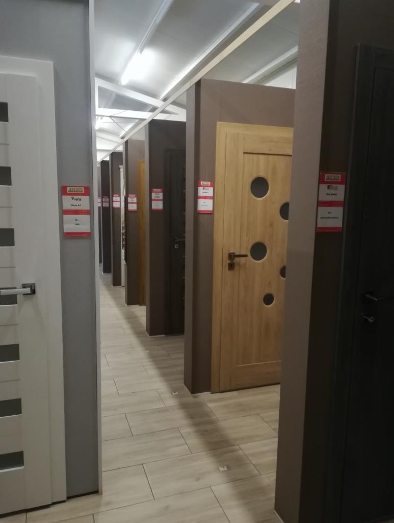 Wystawa rzędu drzwi wewnętrznych o różnych kolorach i wzorach, z etykietami cenowymi, ustawionych wzdłuż korytarza z podłogą imitującą drewno.