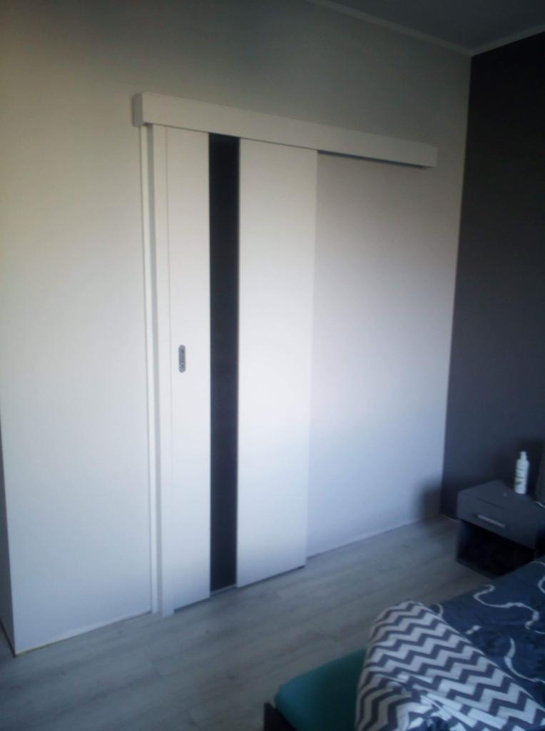 Białe drzwi przesuwne z czarnym pionowym pasem, zamontowane w pokoju z szarą ścianą i jasną podłogą.