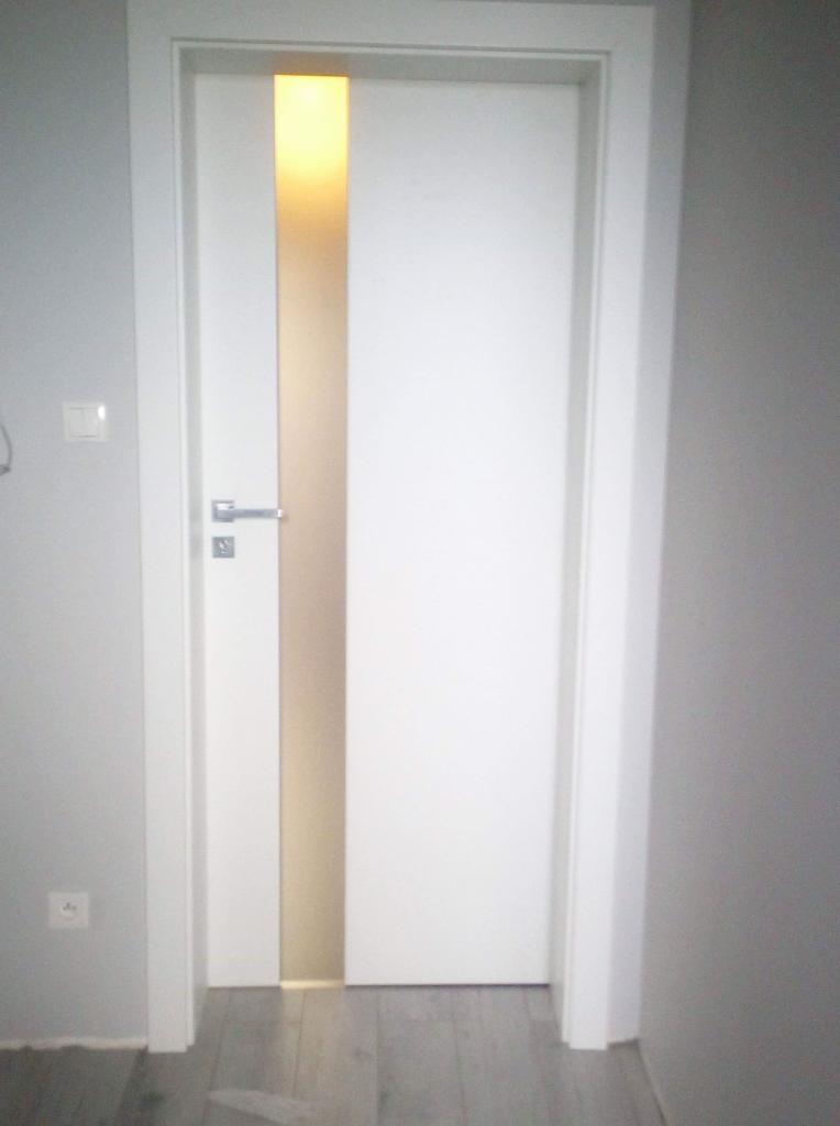 Białe drzwi wewnętrzne z pionowym, podświetlanym paskiem w kolorze ciepłego beżu, zamontowane w szarej ścianie, widoczna klamka i fragment podłogi z paneli.