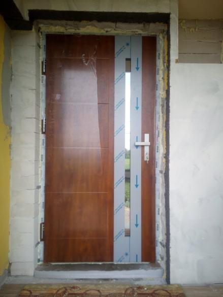 Brązowe drzwi wejściowe z przeszkleniem, w trakcie montażu, widoczne pianka montażowa i folia ochronna na ościeżnicy.