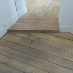 Fragment podłogi z desek po renowacji, widoczny próg z jasnego drewna, białe listwy przypodłogowe, perspektywa skierowana w głąb pomieszczenia.