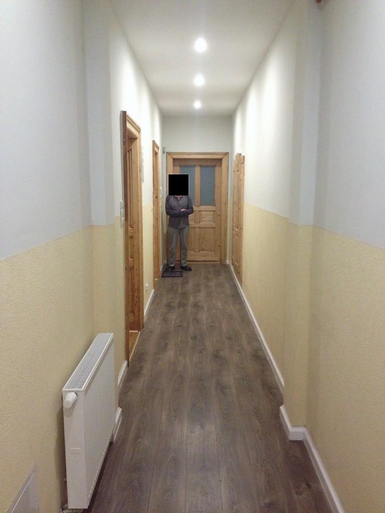 Długi korytarz z drewnianą podłogą i białymi ścianami z żółtym pasem na dole, oświetlony punktowymi lampami sufitowymi; widoczne drzwi z drewnianymi ościeżnicami oraz biały grzejnik przy ścianie.