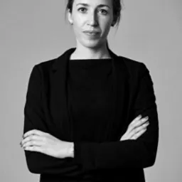 Portret kobiety w czarnym stroju biznesowym, z założonymi rękami, na szarym tle.