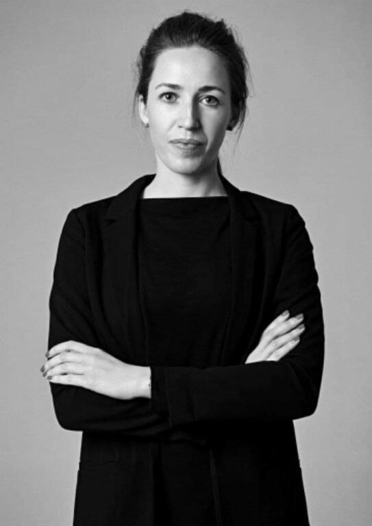 Portret kobiety w czarnym stroju biznesowym, z założonymi rękami, na szarym tle.