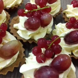 Kruche babeczki z kremem i owocami: ciemne winogrona, czerwone porzeczki, jasne winogrona. Widoczne detale kremu i faktura ciasta.