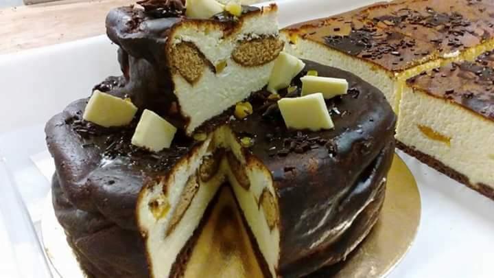 Dwupoziomowy tort z polewą czekoladową, przełożony kremem i biszkoptami, udekorowany kawałkami białej czekolady i posypką czekoladową. W tle widoczny kawałek innego ciasta, prawdopodobnie sernika...