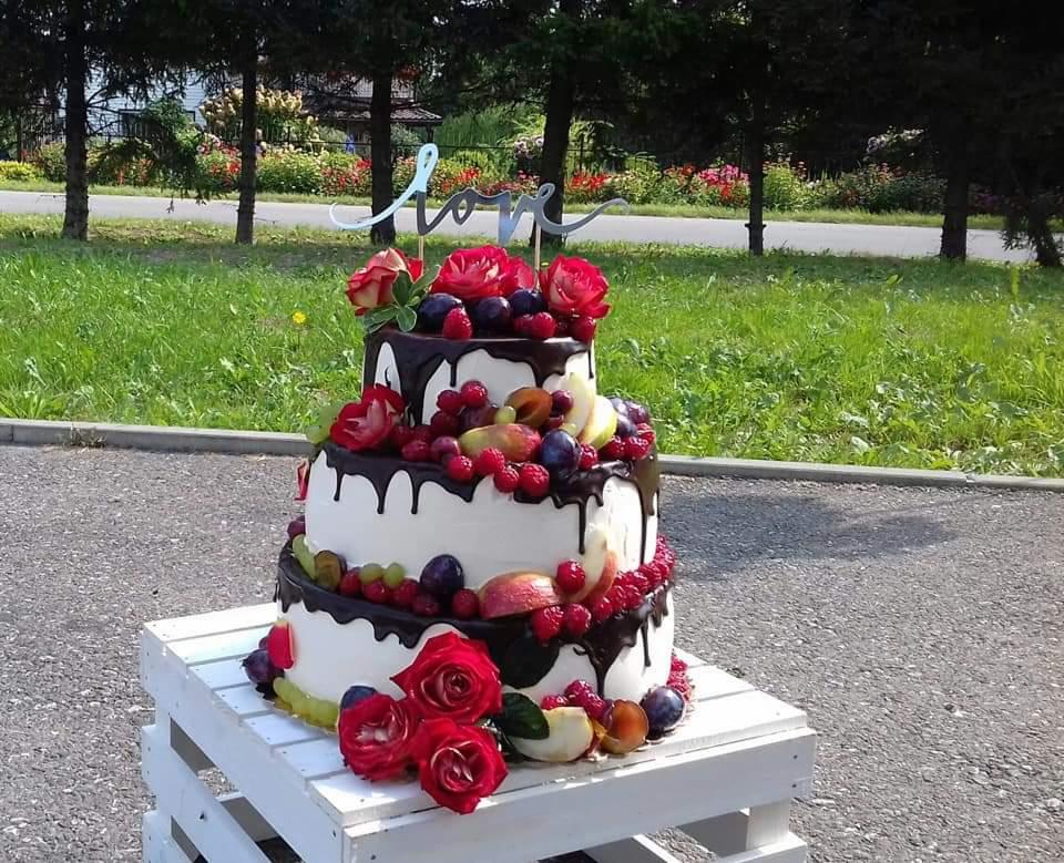 Trzypiętrowy tort weselny z polewą czekoladową, świeżymi owocami i czerwonymi różami, umieszczony na białej, drewnianej skrzynce na tle zielonego trawnika i drzew.