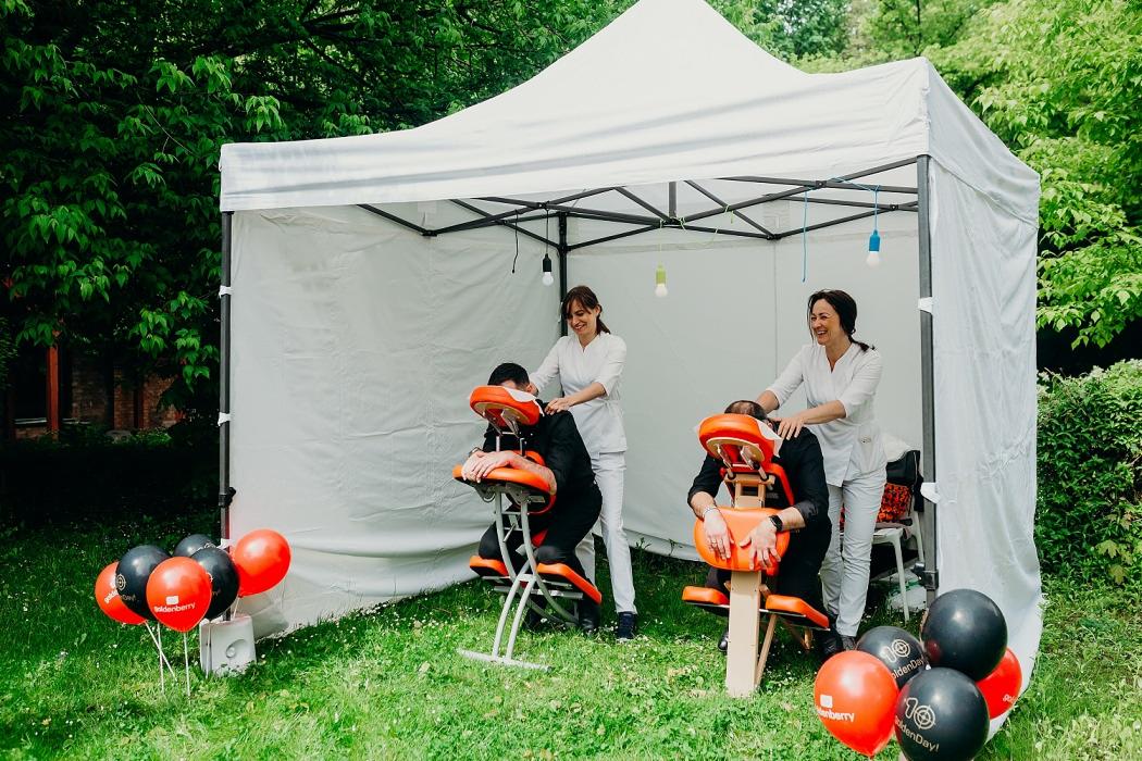 Dwie kobiety w białych uniformach wykonują masaż karku na dwóch mężczyznach siedzących na specjalnych krzesłach do masażu, wszystko pod białym namiotem plenerowym, dekoracja z balonów.