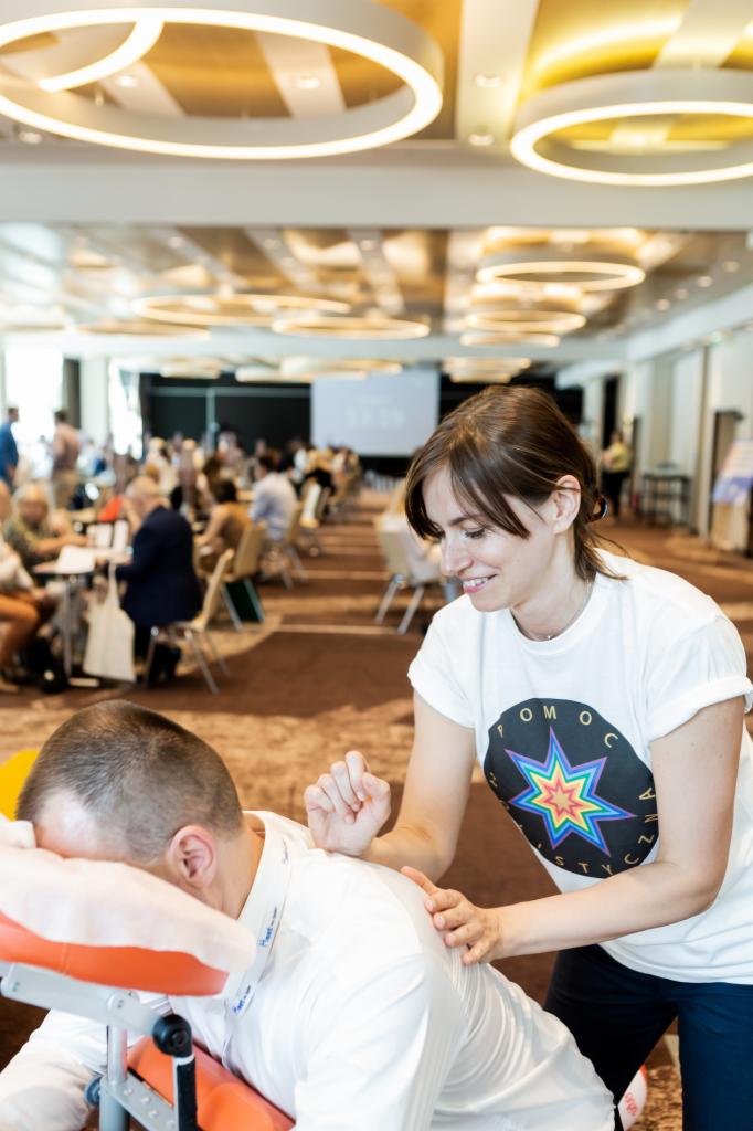 Masażysta w białej koszulce z logo wykonuje masaż pleców mężczyzny na specjalnym krześle do masażu w przestrzeni eventowej, w tle widoczni inni ludzie i stoły.