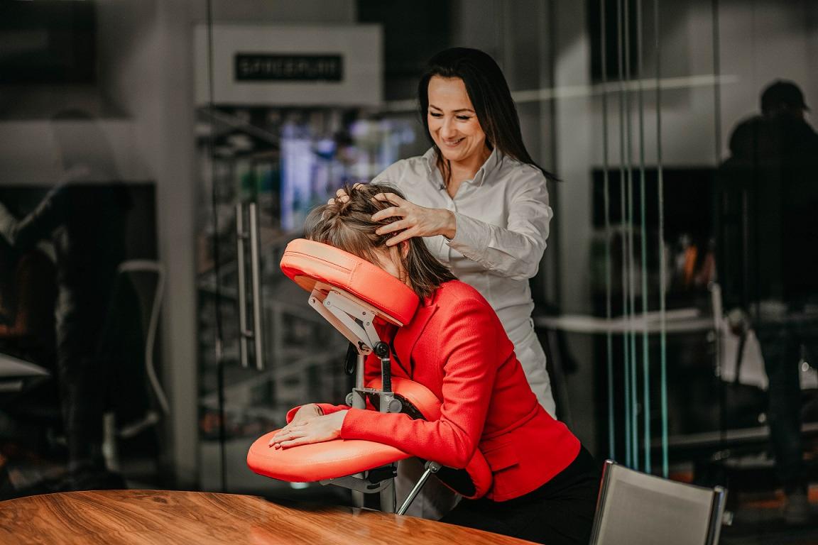 Kobieta w czerwonej marynarce korzysta z masażu głowy i szyi na specjalnym krześle w biurze. Masażystka w białej koszuli delikatnie masuje jej głowę, w tle szklane ściany i biurowy wystrój.