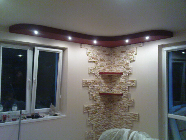 Wykończenie narożnika w salonie z dekoracyjną ścianą z kamienia, podświetlanym sufitem i półkami, widok na okno z parapetem zastawionym przedmiotami.