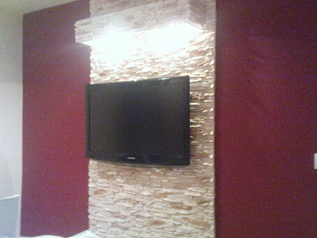 Telewizor zawieszony na ścianie z dekoracyjną okładziną imitującą kamień, obok ściana pomalowana na bordowo, realizacja w Sosnowcu.