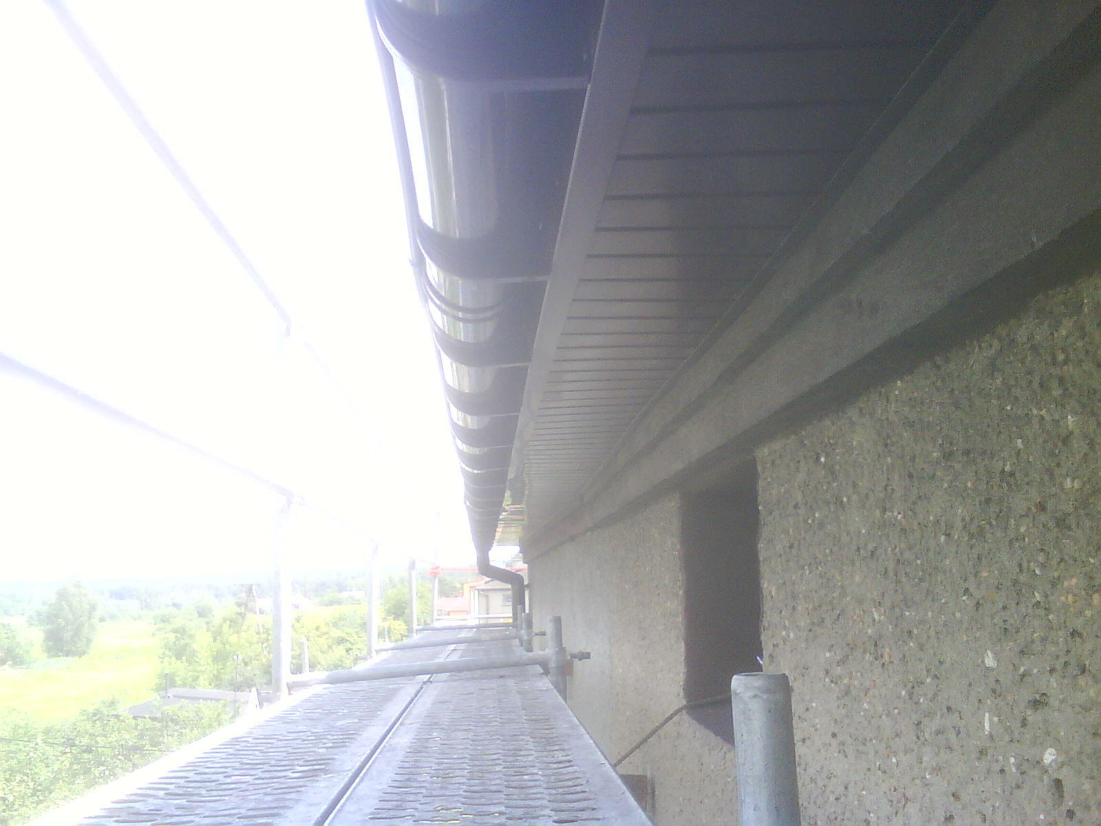 Widok z rusztowania na zamontowany system rynnowy przy budynku z elewacją z kamyczków, w tle zieleń drzew.