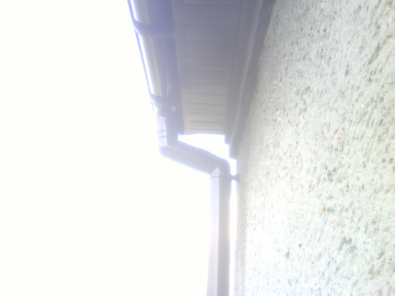 Szary system rynnowy zamontowany przy szorstkiej, jasnej ścianie budynku, widoczne elementy mocujące.