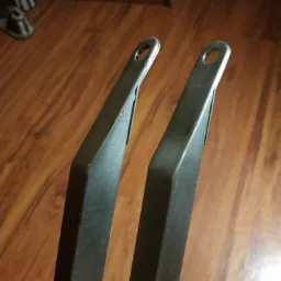 Dwa metalowe elementy pomalowane proszkowo na czarno, z otworami na końcach, leżące na drewnianej podłodze.