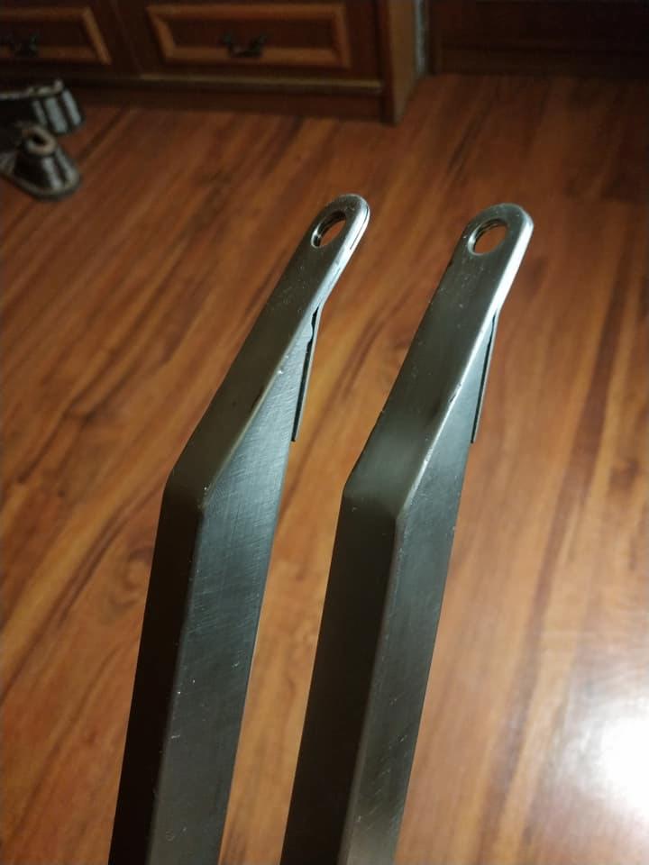 Dwa metalowe elementy pomalowane proszkowo na czarno, z otworami na końcach, leżące na drewnianej podłodze.