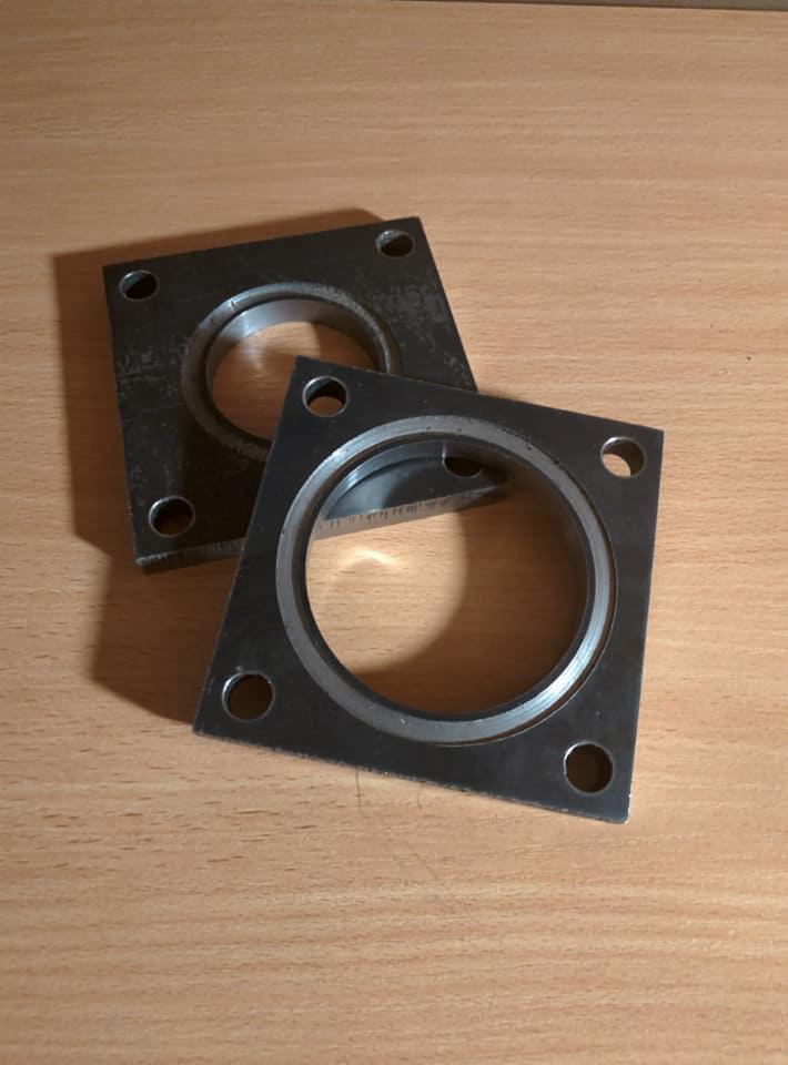 Dwie metalowe, kwadratowe płyty z centralnym okrągłym otworem i czterema otworami montażowymi w rogach, leżące na drewnianej powierzchni.