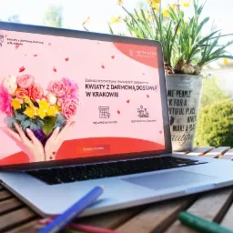 Laptop wyświetlający stronę internetową z ofertą kwiatów z darmową dostawą w Krakowie, na drewnianym stole, obok doniczki z żonkilami i kredek.