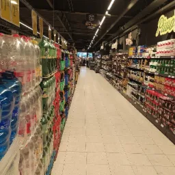 Wnętrze sklepu Netto: widok na alejkę z napojami w plastikowych butelkach, oświetloną podwieszanymi lampami liniowymi, z widocznymi cenami na górnych półkach i dekoracyjnymi napisami na ścianach.