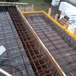 Zbrojenie fundamentów w szalunku z żółtymi elementami, widoczne bloczki Solbet w tle, oraz narzędzia YATO.