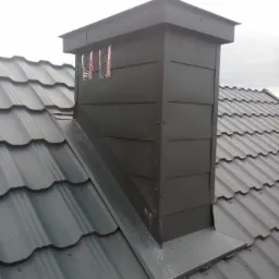 Metalowy komin z wentylacją na dachu pokrytym blachodachówką w deszczowy dzień.