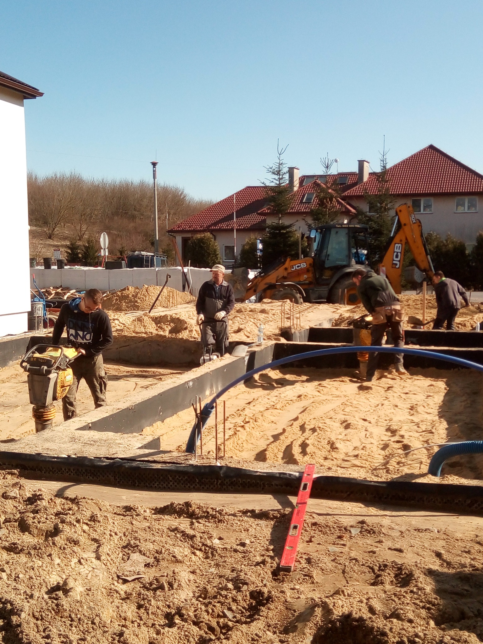 Prace ziemne pod fundamenty domu: zagęszczanie gruntu ubijarką, widoczny wykop, szalunki, zbrojenie, poziomica i koparka JCB w tle.