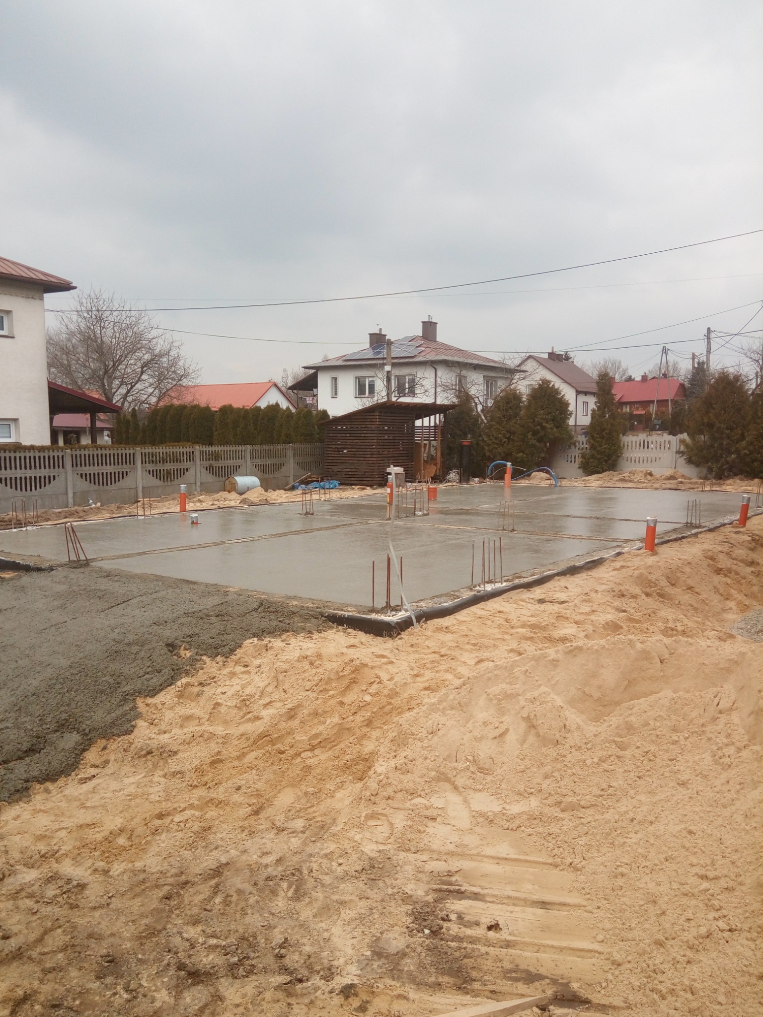 Wylana, świeża płyta fundamentowa na piaszczystym podłożu, z wystającymi prętami zbrojeniowymi, pomarańczowe rury instalacyjne, w tle zabudowa jednorodzinna pod pochmurnym niebem.