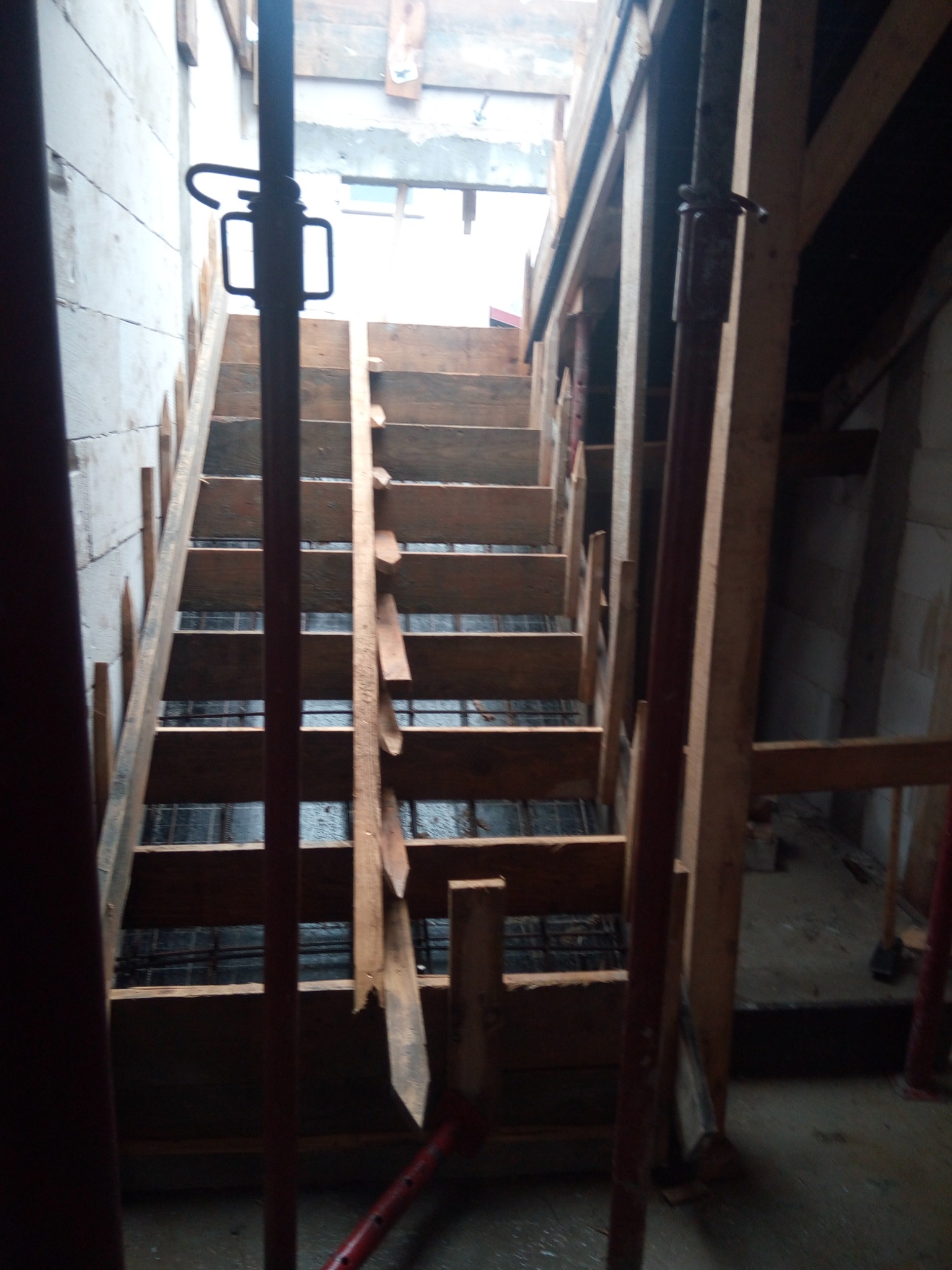 Szczegółowe ujęcie drewnianego szalunku schodów w trakcie budowy, z widocznym zbrojeniem i podporami stalowymi, oświetlonym przez światło wpadające z góry.