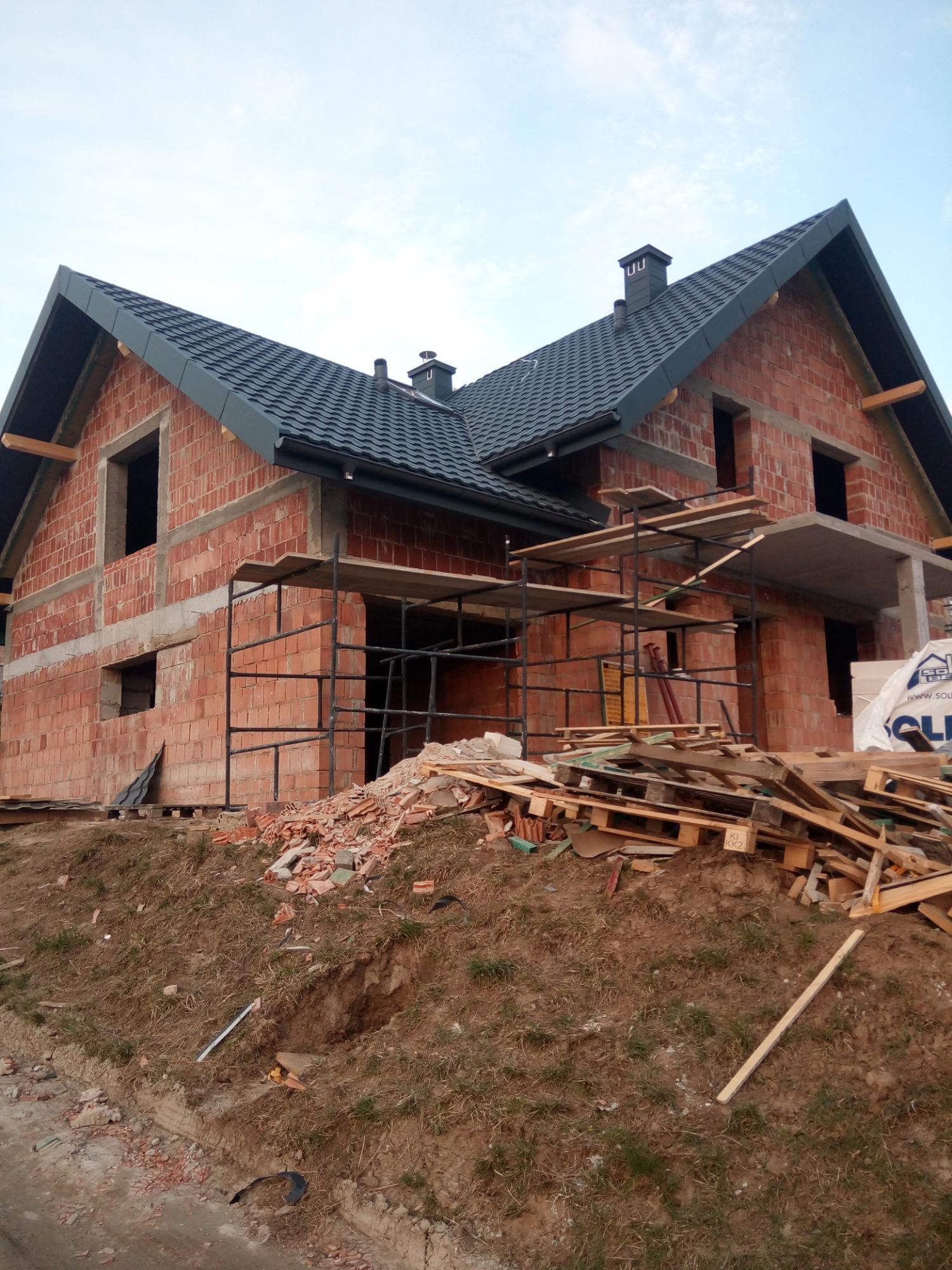 Surowy, dwupiętrowy budynek w trakcie budowy z czerwonej cegły, z zamontowanym ciemnoszarym dachem z dachówki, otoczony rusztowaniami, paletami i gruzem.