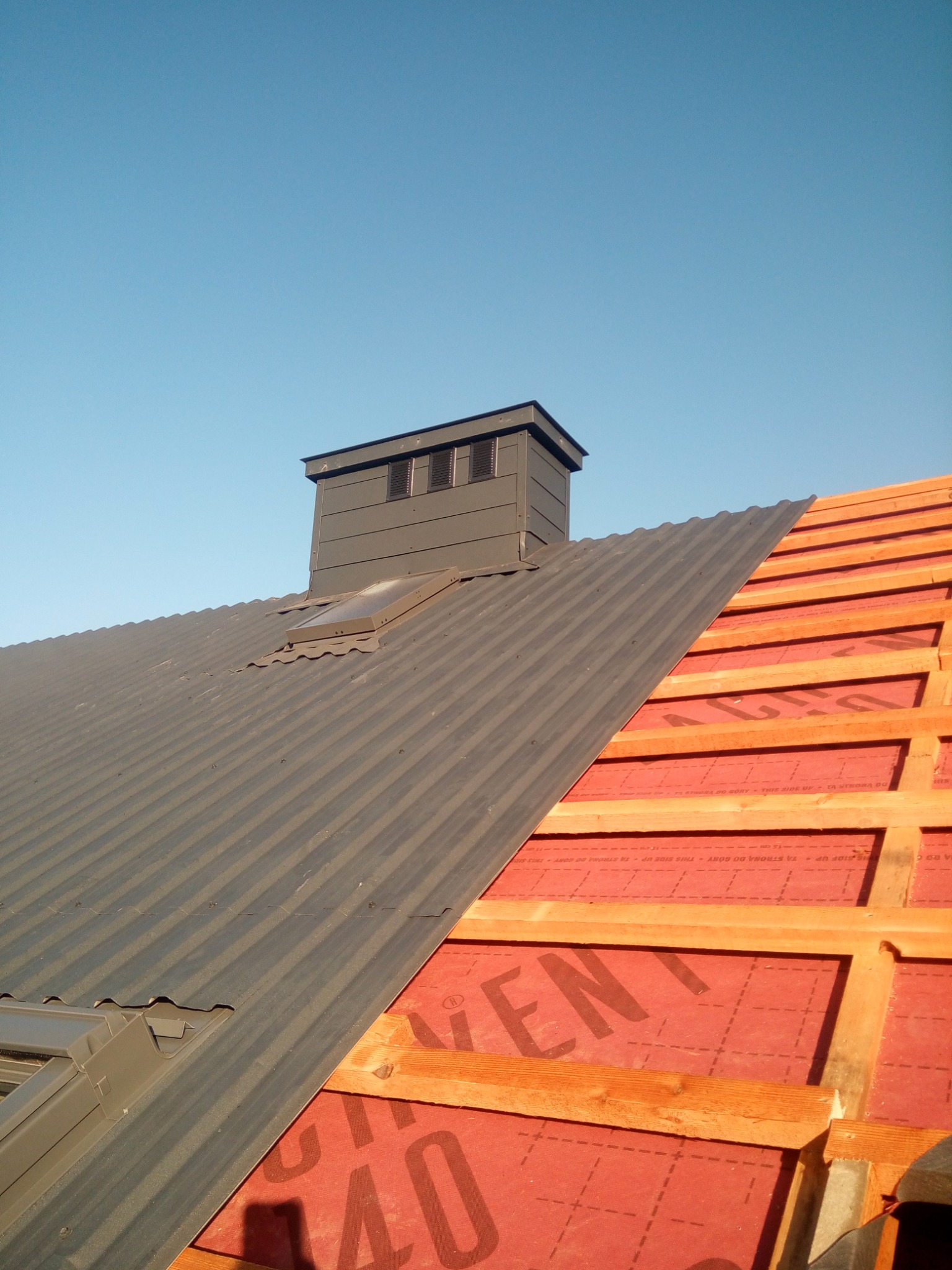 Częściowo pokryty blachą dach z kominem i oknem dachowym, widoczna więźba dachowa z czerwoną membraną w trakcie prac.