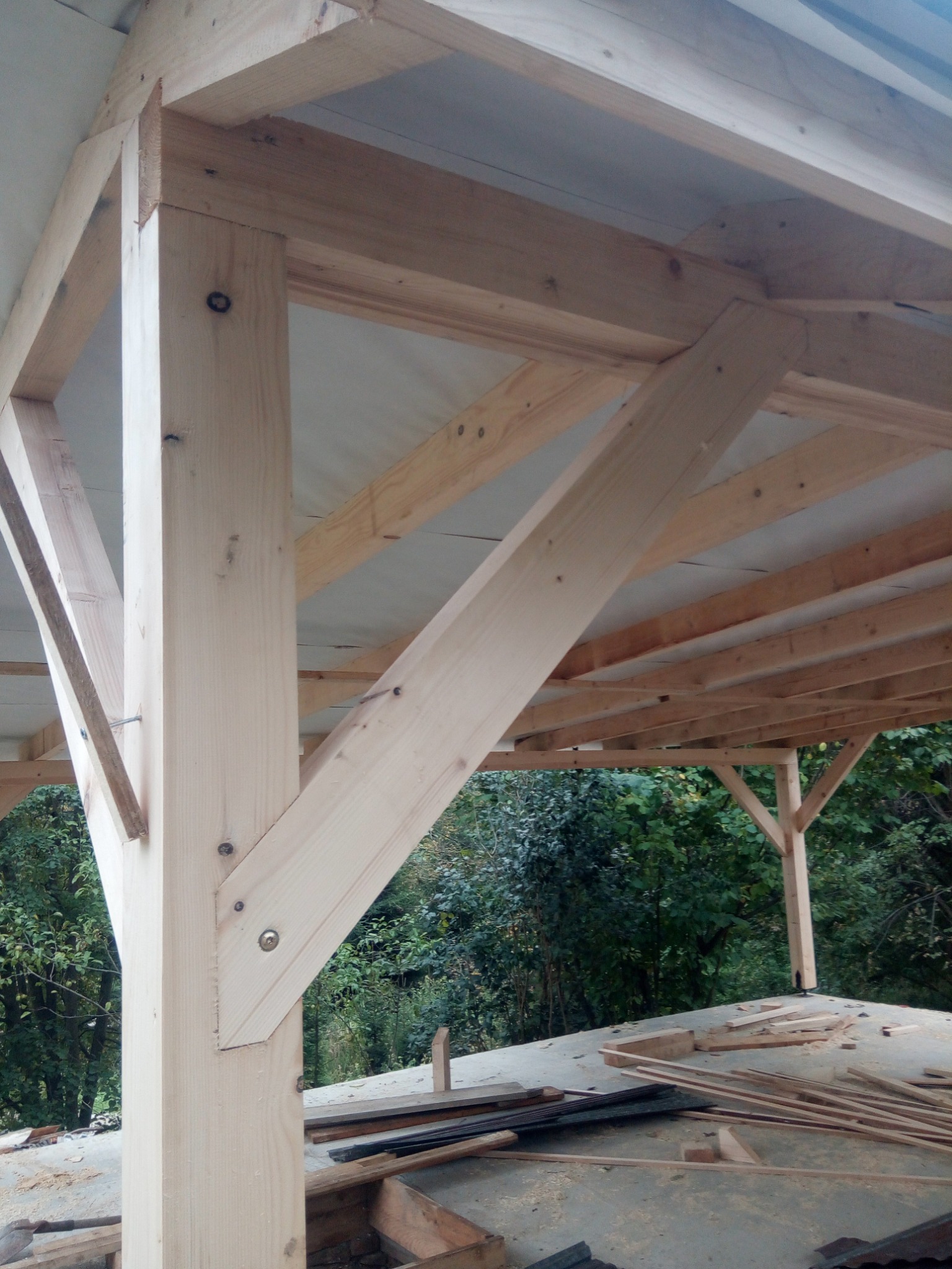 Konstrukcja szkieletowa z jasnego drewna w trakcie montażu, widoczne połączenia śrubowe i fragment poszycia dachu, w tle zieleń drzew.
