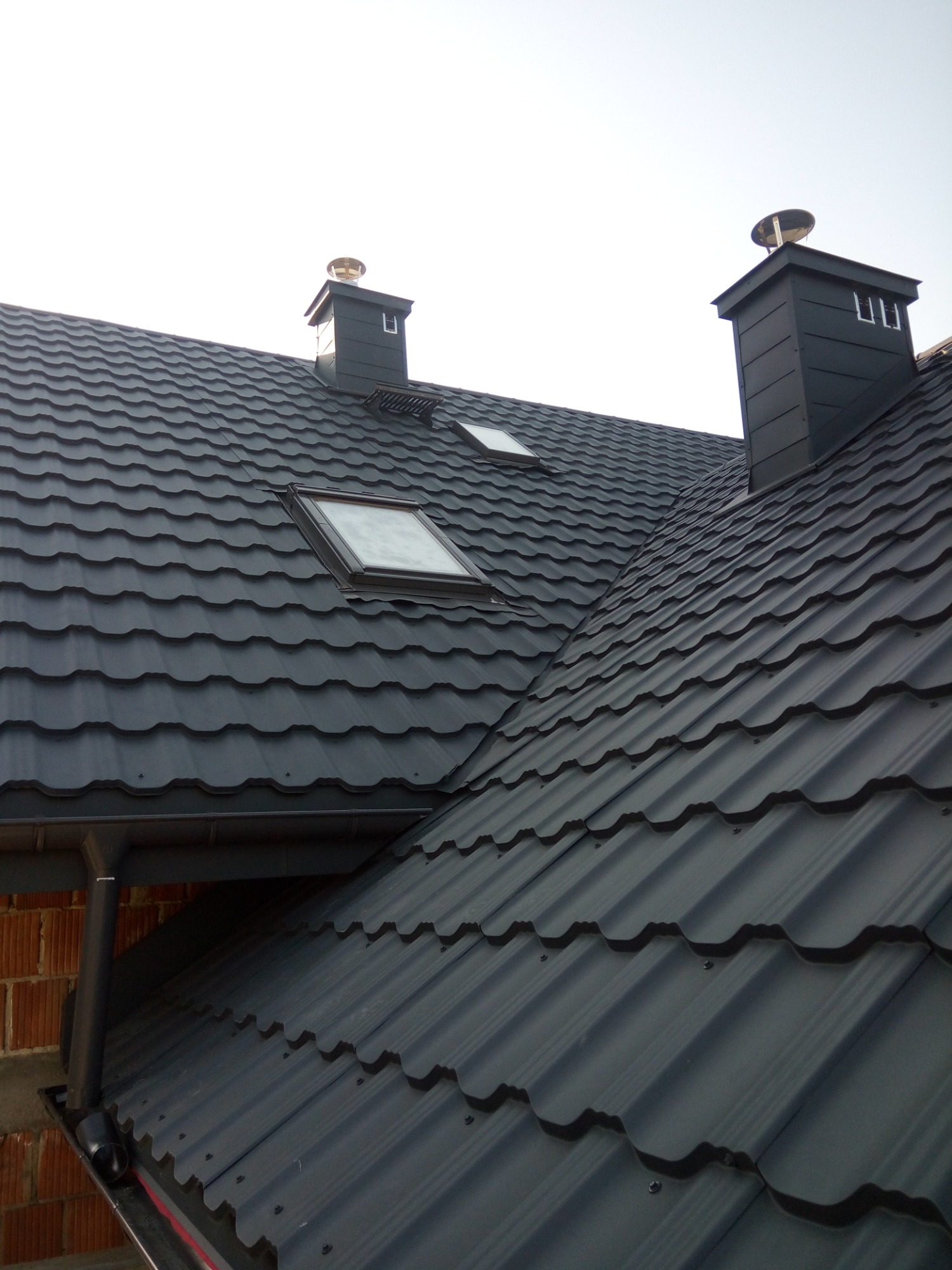 Nowoczesny, grafitowy dach z blachodachówki trapezowej, z dwoma oknami dachowymi i dwoma kominami z metalowymi nasadami, widok z dołu na tle jasnego nieba.