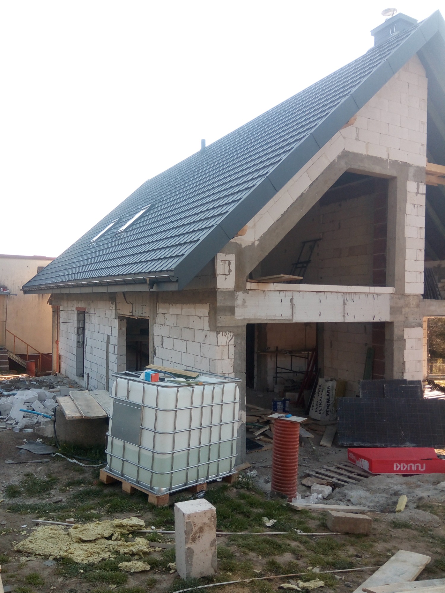 Niedokończony, dwukondygnacyjny budynek z białego bloczka z szarym dachem, widoczne okno dachowe, fragment poddasza z niezabezpieczonym otworem, na pierwszym planie zbiornik IBC, materiały...