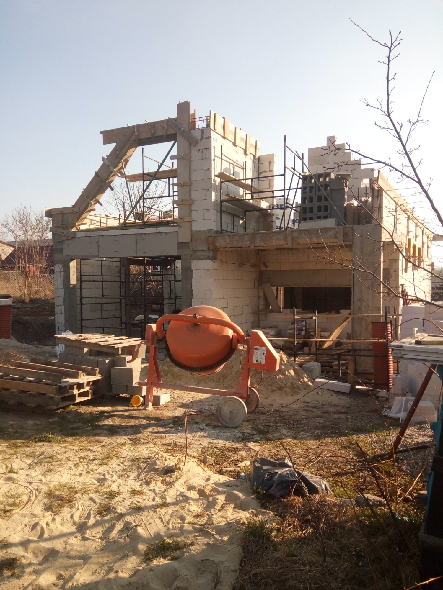 Wznoszenie ścian domu murowanego z betonu komórkowego, widoczna pomarańczowa betoniarka, drewniane szalunki i zbrojenia, w tle zabudowa jednorodzinna i błękitne niebo.