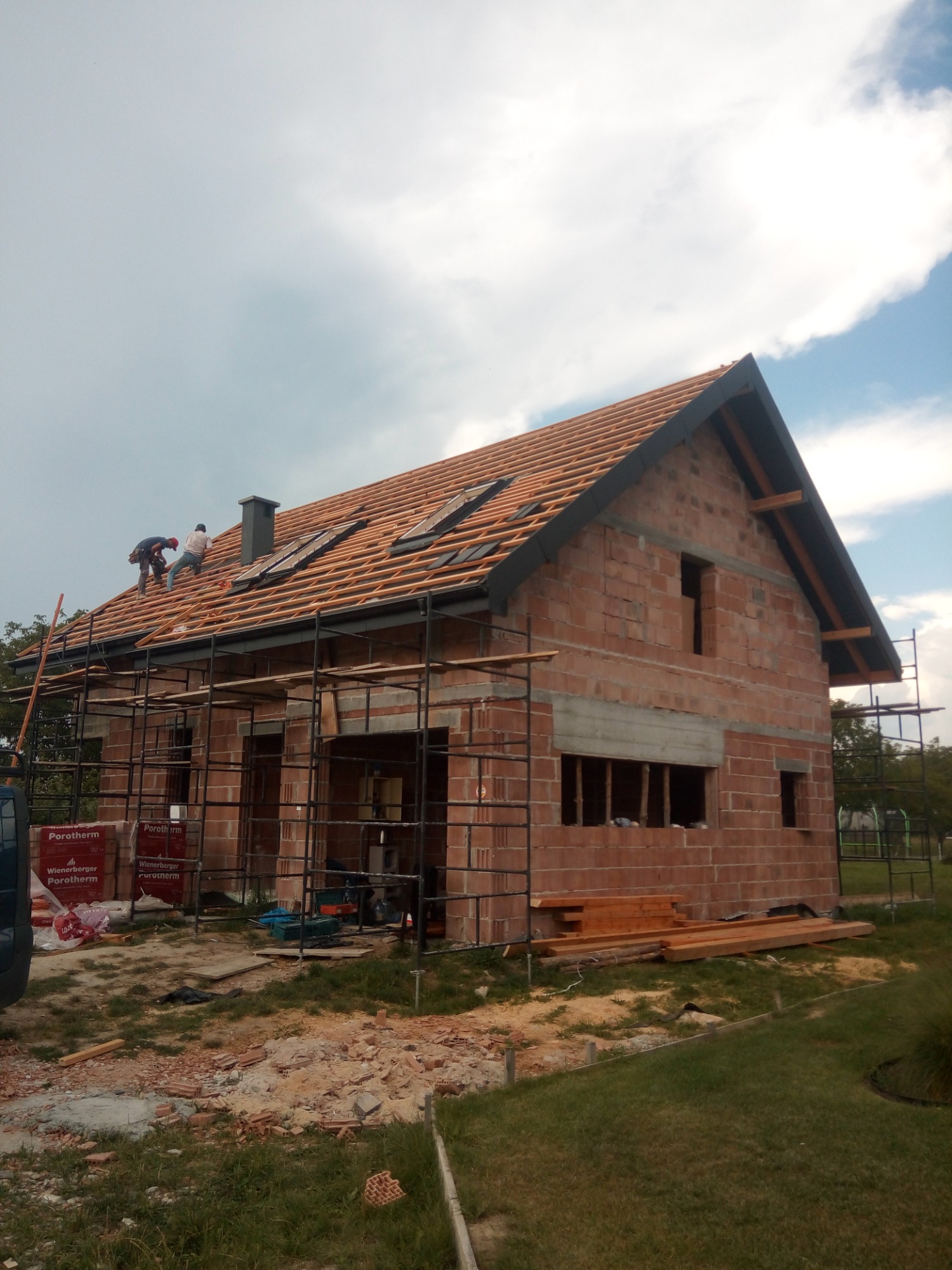 Budowa domu z cegły Porotherm: dekarze na rusztowaniu montują drewnianą konstrukcję dachu z oknami dachowymi, widoczne materiały budowlane i narzędzia.