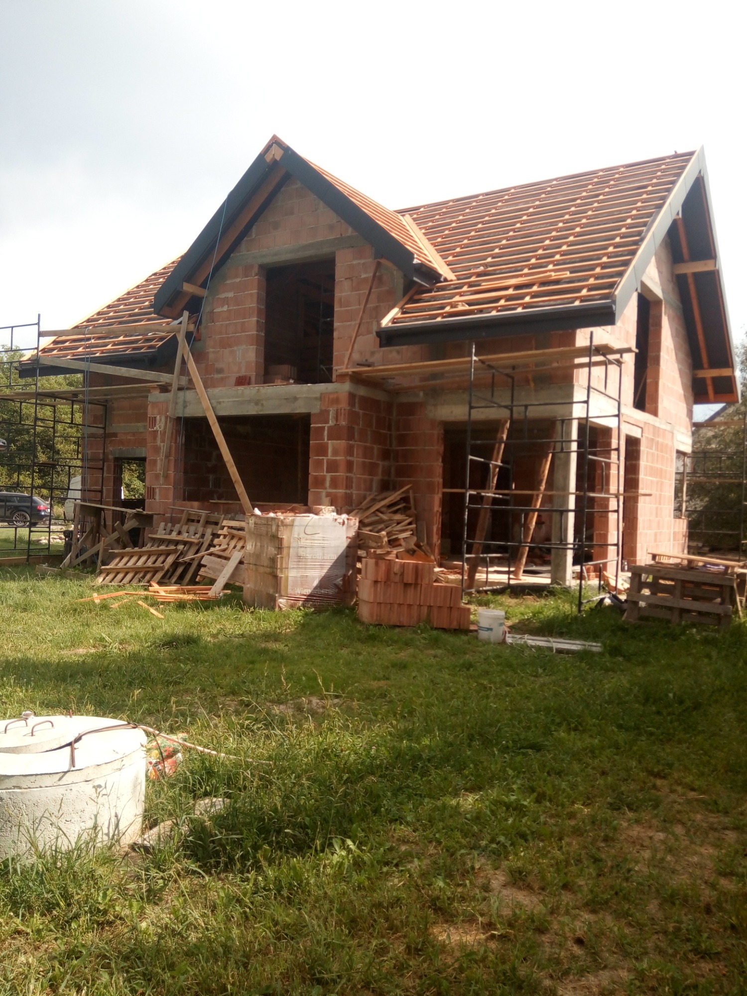 Niedokończony murowany dom z częściowo ułożoną konstrukcją dachu, widoczne rusztowania i składowane materiały budowlane na trawniku.