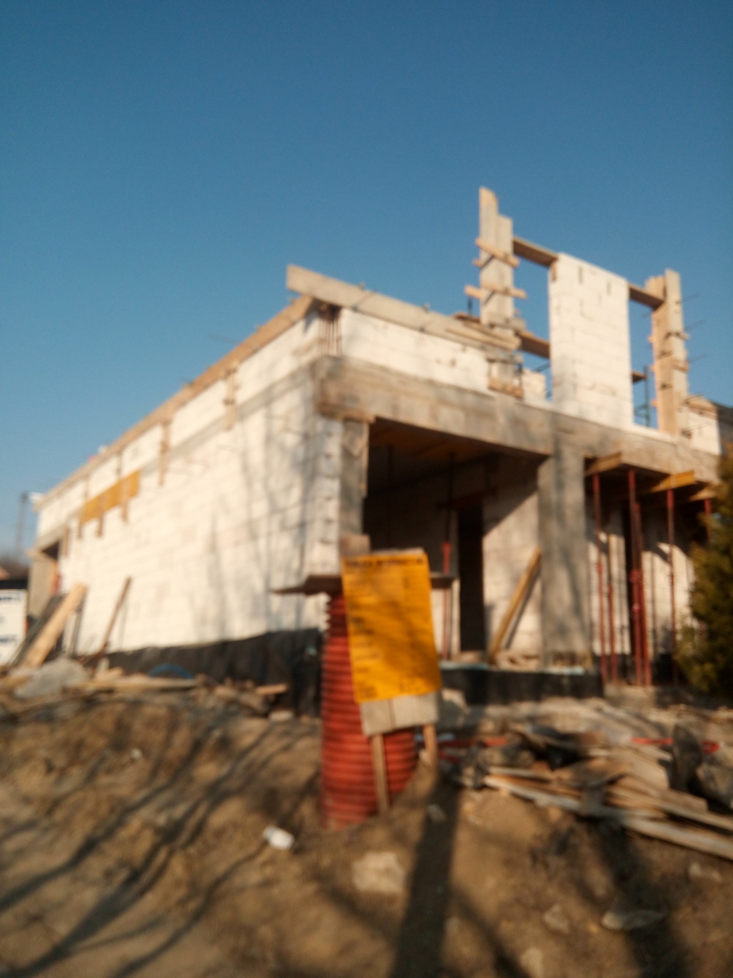 Wznoszony dwukondygnacyjny budynek z białych bloczków, widoczne drewniane szalunki, metalowe podpory i żółta tablica informacyjna na placu budowy w słoneczny dzień.