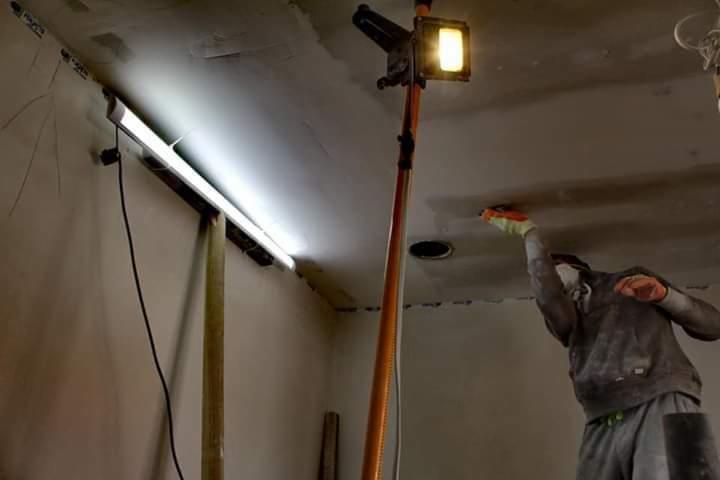 Pracownik w masce i rękawicach szpachluje sufit w pomieszczeniu oświetlonym lampą budowlaną i lampą LED.