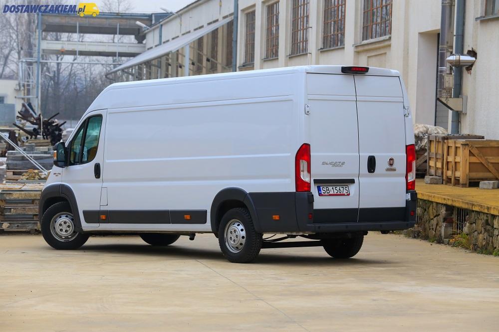 Biały samochód dostawczy Fiat Ducato z widoczną tablicą rejestracyjną, zaparkowany na betonowym placu przed budynkiem przemysłowym.