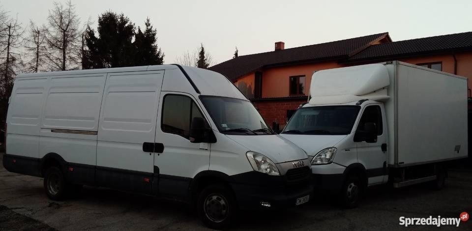 Dwa białe busy transportowe marki Iveco zaparkowane blisko siebie na tle budynku mieszkalnego i drzew, sfotografowane o zmierzchu.