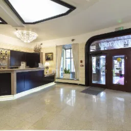 Eleganckie lobby hotelowe z recepcją, kryształowym żyrandolem i przeszklonymi drzwiami wejściowymi z napisem 'Victoria Hotel'. Na ścianie zegar i dekoracje.