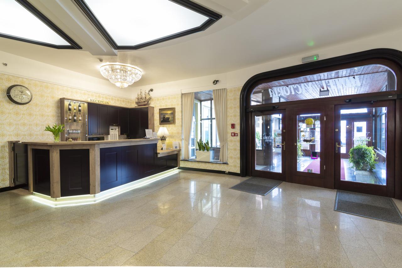 Eleganckie lobby hotelowe z recepcją, kryształowym żyrandolem i przeszklonymi drzwiami wejściowymi z napisem 'Victoria Hotel'. Na ścianie zegar i dekoracje.