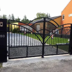 Czarna, metalowa brama wjazdowa z numerem 193, ozdobiona łukami i kratą, na tle żwirowego podjazdu i zieleni ogrodu. Widoczny dom z pomarańczową elewacją.