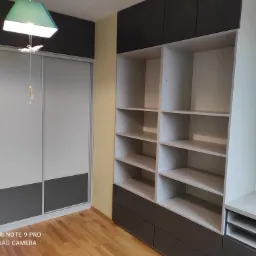 Wnętrze pokoju z zabudową meblową: biało-szara szafa przesuwna z dekoracyjnym oświetleniem sufitowym oraz regał z szufladami w tej samej kolorystyce, na drewnianej podłodze.