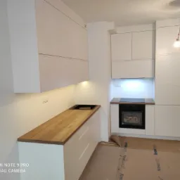 Nowoczesna, minimalistyczna kuchnia z białymi szafkami i drewnianym blatem, widok na piekarnik i niezabezpieczoną żarówkę zwisającą z sufitu.